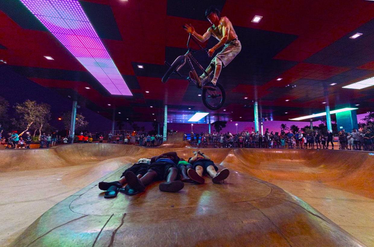 A BMX rider performing an aerial stunt, jumping over three people lying on the ground in a skate park illuminated by LED pixel dot lights.